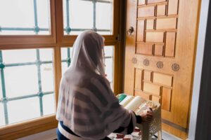 A woman in a headscarf is praying inside a mosque, reading the Quran by a window.